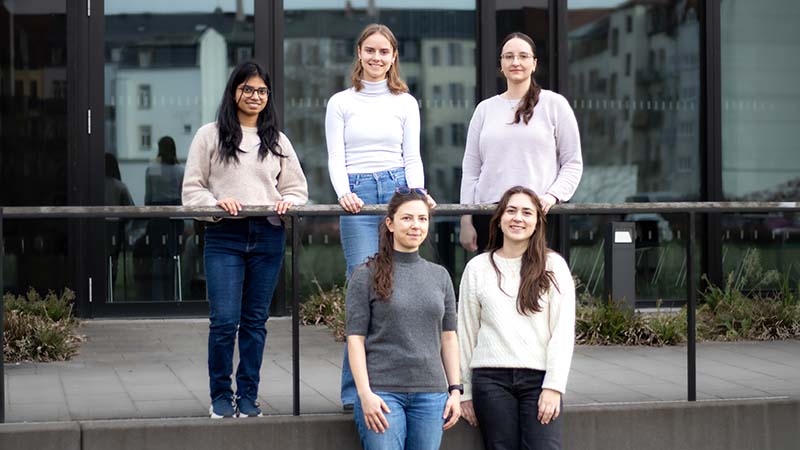 Five young people in front of a dark building with lots of glass. Three of them standing on a raised level behind a handrail, two standing lower before the raised level
